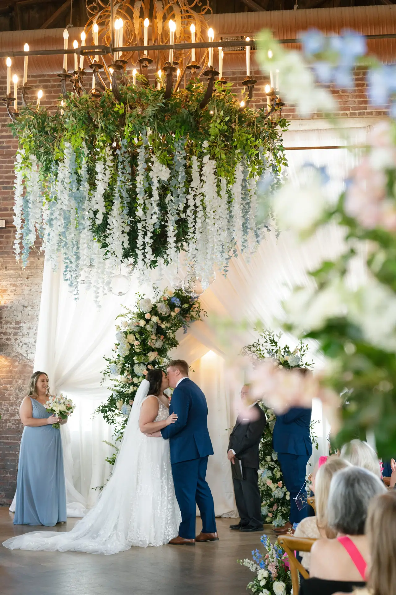 Bride and groom share their first kiss under a floral arch with cascading greenery and white drapery.