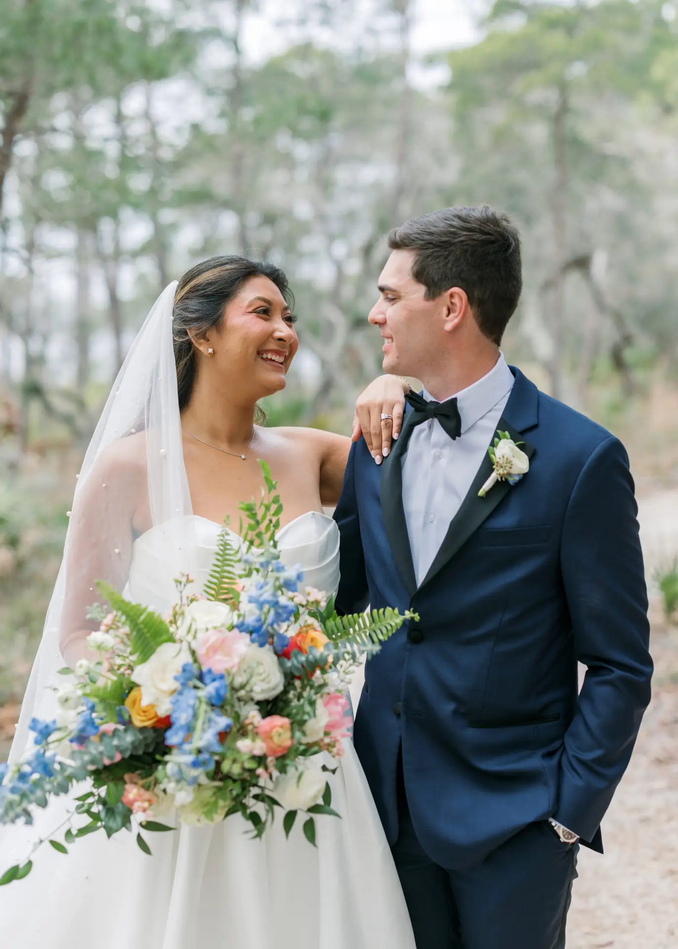 Bride and groom smiling outdoors in forest setting, bride holding bouquet of pastel and blue flowers.
