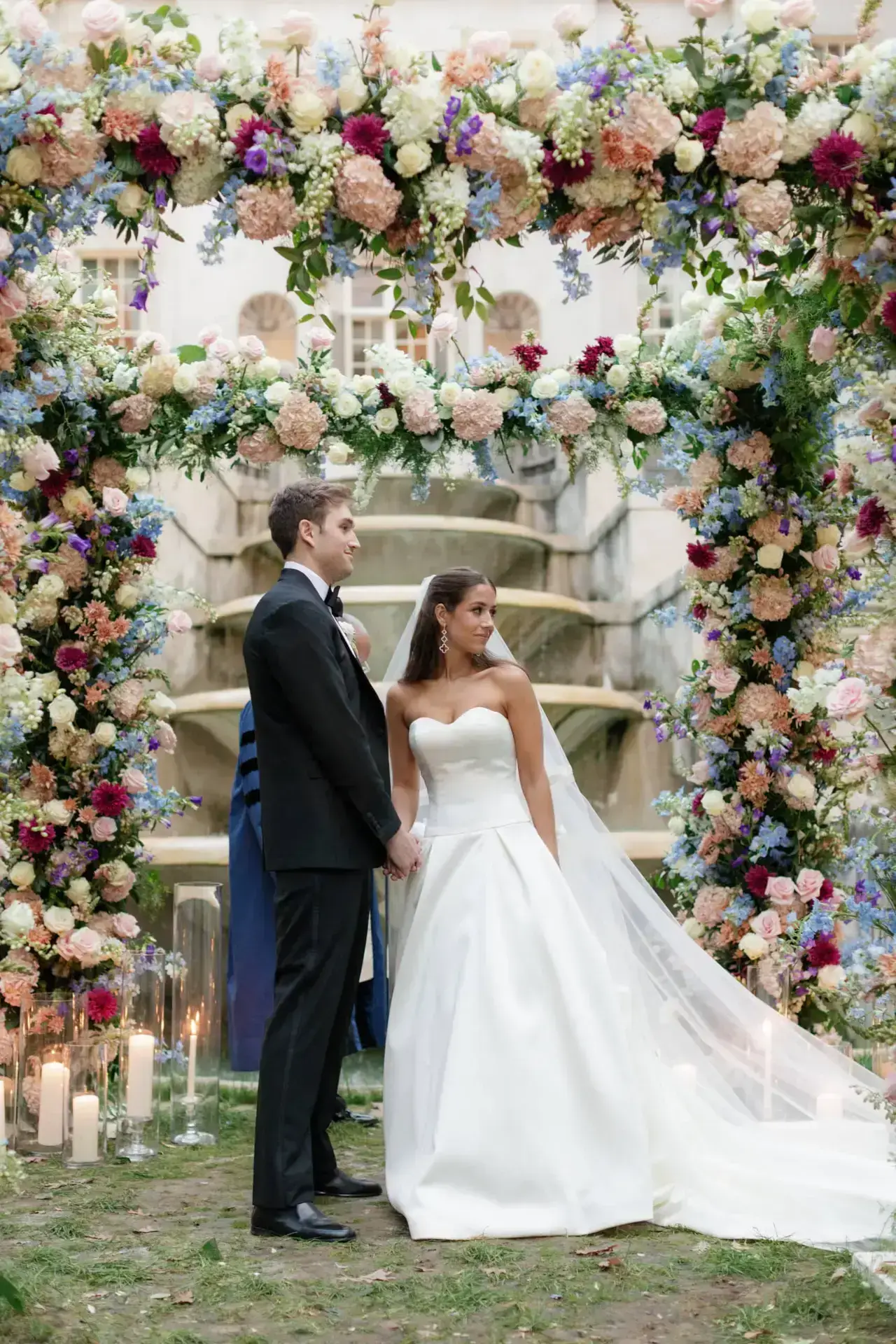 Bride and groom standing under a lush floral arch with pastel roses, hydrangeas, and candles during outdoor wedding ceremony.