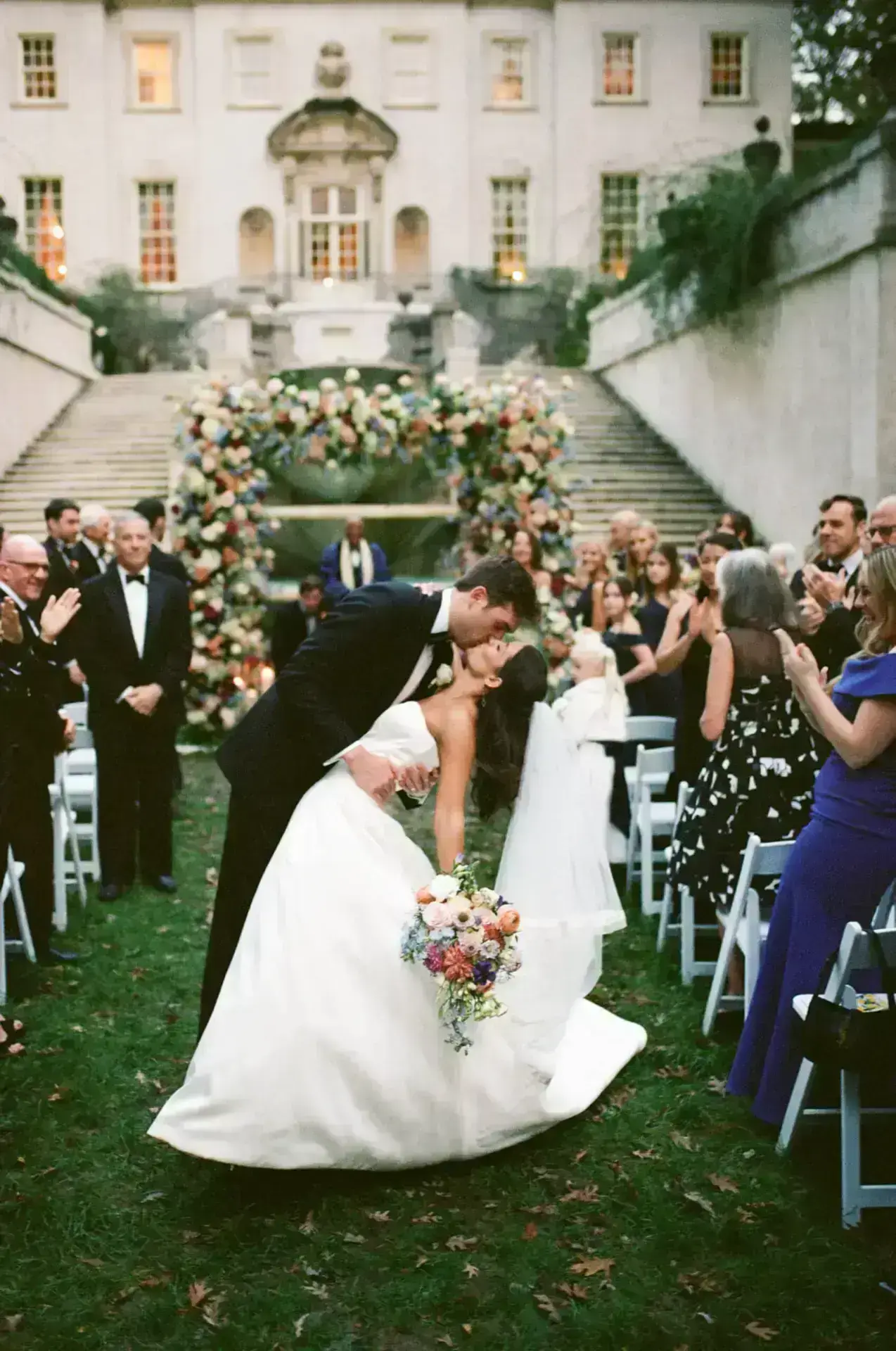 Groom dips bride for a romantic kiss surrounded by guests applauding at elegant outdoor wedding ceremony.