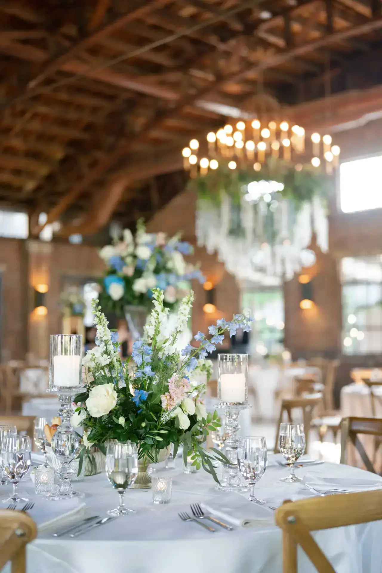Wedding reception table decorated with tall blue and white floral centerpiece, candles, and elegant tableware.